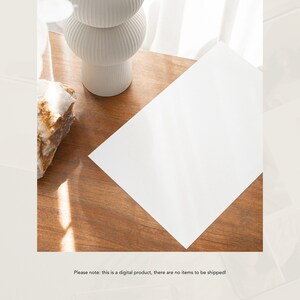 May include: A white ceramic vase with a textured design sits on a wooden table with a white sheet of paper on top. The paper is blank and the table is illuminated by sunlight.