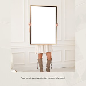 May include: A woman is holding a large, blank canvas in a gold frame in front of her. The canvas is white and the frame is a thin, gold metal. The woman is wearing a white dress and brown boots. The background is a white wall with white trim.