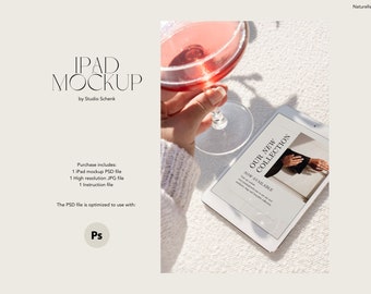 May include: A white iPad mockup with a screen displaying a graphic design for a new collection. The text on the screen reads "Our New Collection Now Available". The iPad is on a white textured surface with a glass of pink liquid in the background.