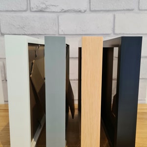 Pode incluir: Quatro molduras de fotos em cores diferentes: branco, cinza, madeira clara e preto. As molduras s&atilde;o todas do mesmo tamanho e est&atilde;o alinhadas uma ao lado da outra.