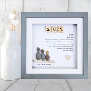 Pode incluir: Uma pe&ccedil;a de arte de seixos enquadrada com a palavra "SON" escrita em pe&ccedil;as de Scrabble. A arte de seixos representa uma fam&iacute;lia de tr&ecirc;s, com o texto "Dad, Mum, Harry" abaixo da arte. A moldura &eacute; cinza e o fundo &eacute; branco. O texto abaixo da arte de seixos diz: "Um menino que &eacute; a menina dos olhos de sua m&atilde;e, o maior orgulho de um pai. Um barril barulhento de risos. Descarado, corajoso, engra&ccedil;ado e forte. Algu&eacute;m que segura sua m&atilde;o por um tempo, mas que guarda seu cora&ccedil;&atilde;o para sempre. Veja tamb&eacute;m: b&ecirc;n&ccedil;&atilde;o."