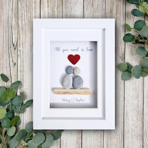 May include: A framed artwork with a white border. The artwork features a red heart above two pebble figures, with the text "All you need is love." Below, the names "Harry & Sophie" are written. The background is white.