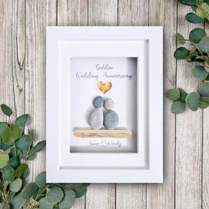 May include: A framed art piece for a golden wedding anniversary. The white frame surrounds a design with pebble figures, a gold heart, and the words "Golden Wedding Anniversary." The names "James & Wendy" are also included.