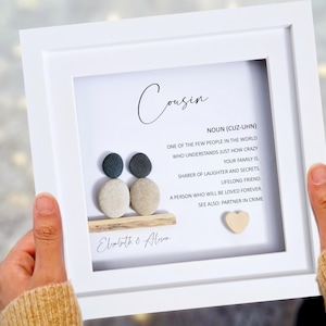 May include: White framed picture with a pebble art design of two people sitting on a driftwood log. The text reads "Cousin, Noun (Cuz-Uhn) One of the few people in the world who understands just how crazy your family is. Sharer of laughter and secrets. Lifelong friend. A person who will be loved forever. See also: Partner in crime. Elizabeth & Alison"