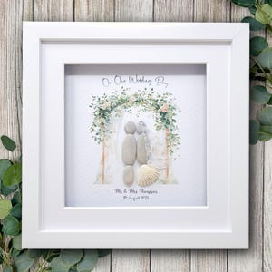 May include: A framed pebble art piece with a wedding theme. The artwork features pebble figures under a floral arch, with the text "On Our Wedding Day" and a personalized name and date. The frame is white.