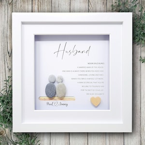 May include: A white framed artwork with the word "Husband" written in script. The artwork includes a definition of the word, pebble art of a couple, and the names "Paul & Jenny". A small wooden heart is also included.