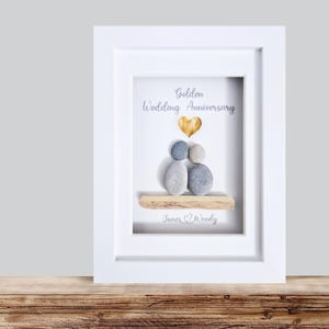May include: A framed art piece for a golden wedding anniversary. The artwork features pebble figures, a gold heart, and the words "Golden Wedding Anniversary" and a couple's names. The frame is white, and the background is light.