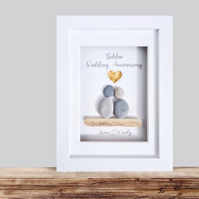 May include: A framed art piece for a golden wedding anniversary. The artwork features pebble figures, a gold heart, and the words "Golden Wedding Anniversary" and a couple's names. The frame is white, and the background is light.
