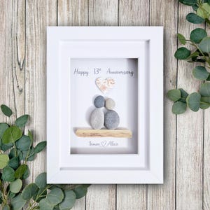 May include: A framed artwork with a white border. The artwork features a pebble art design with two figures, a heart, and the text "Happy 13th Anniversary." The names "Simon & Alice" are also included. The background is a light color.