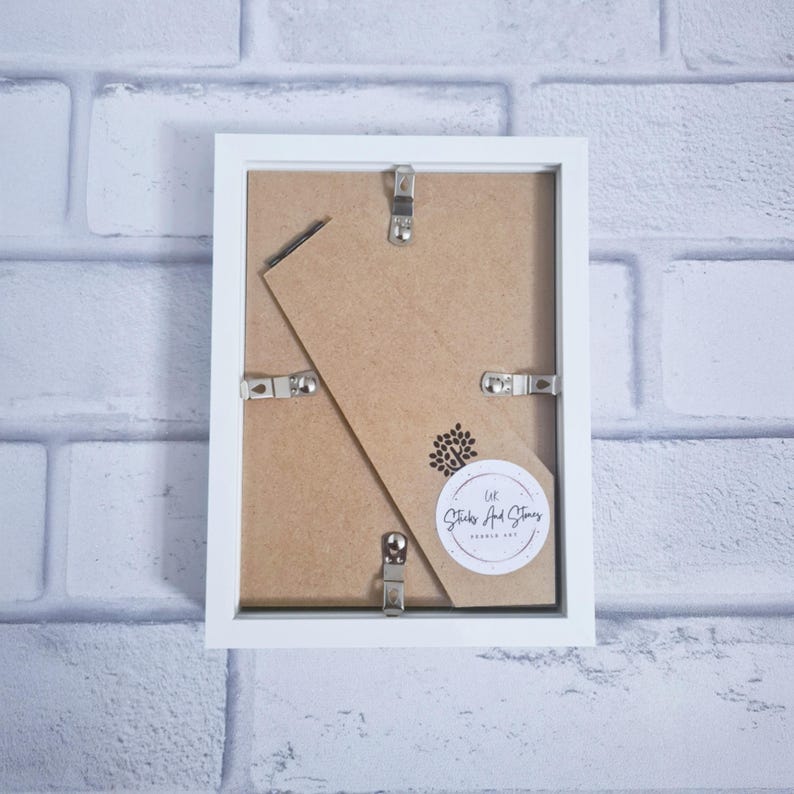 May include: Back of a white picture frame with metal clips and a cardboard backing. A small, round sticker with a tree design and text is visible. The frame is against a white brick wall.