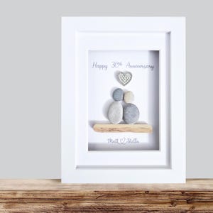 May include: A framed pebble art piece with a white frame. The art features two pebble figures, a heart, and the text "Happy 30th Anniversary" and "Matt & Stella". The art is set on a wooden base.