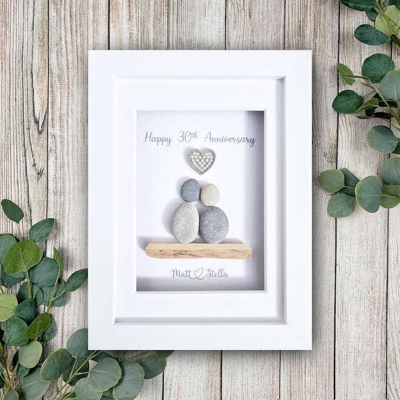 May include: White framed art piece with a pebble art design. The art features two pebble figures, a heart, and the text "Happy 30th Anniversary" and "Matt & Stella". The background is white, and the frame is white.