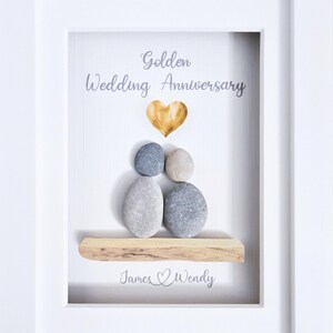 May include: Framed artwork for a golden wedding anniversary. The white frame surrounds a white background with the words "Golden Wedding Anniversary" and a gold heart. Pebble art depicts a couple, with the names "James & Wendy" below.