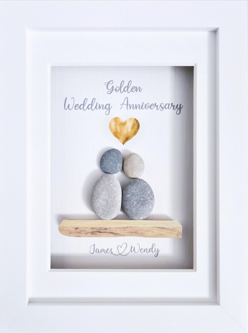 May include: Framed artwork for a golden wedding anniversary. The white frame surrounds a white background with the words "Golden Wedding Anniversary" and a gold heart. Pebble art depicts a couple, with the names "James & Wendy" below.