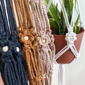 May include: Close-up of several macrame plant hangers in various colors: navy, brown, tan, and white. Each hanger features a flower-shaped detail with a wooden bead center. A potted plant with green and yellow leaves is visible in one of the white hangers.