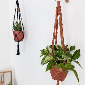 May include: Two macrame plant hangers with wooden beads hanging from a wall. The hanger on the left is black and the hanger on the right is terracotta. Both hangers have a crescent moon-shaped hook.