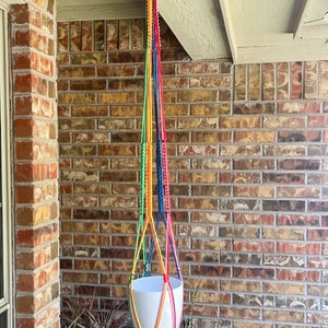 May include: A rainbow macrame plant hanger with a white ceramic pot hanging from a hook on a brick wall.