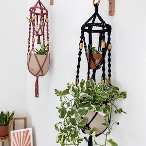 May include: Two macrame plant hangers with wooden beads, one in black and one in pink, hanging from wooden brackets on a white wall. Both hangers have a pot with a plant inside.