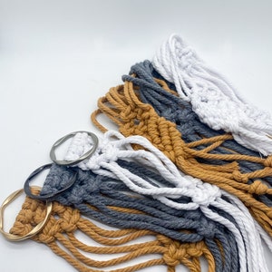 May include: Three macrame plant hangers with gold, gray, and white cords. The hangers are attached to metal rings.