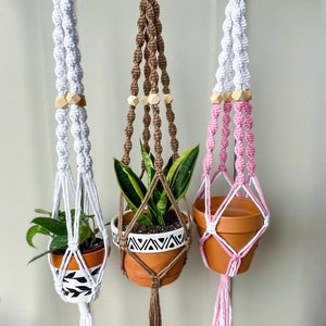 May include: Three macrame plant hangers with wooden beads. The hangers are white, brown, and pink. Each hanger has a small potted plant inside.