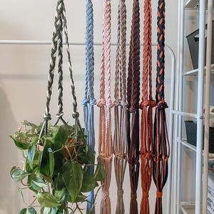 May include: A collection of handmade macrame plant hangers in various colors, including green, blue, pink, and brown. Each hanger features a wooden ring at the top and a braided design. One hanger holds a potted plant with green leaves.