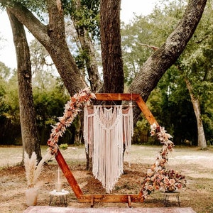 May include: A wooden hexagonal archway decorated with dried flowers and a macrame wall hanging. The archway is set up outdoors in a grassy area with trees in the background.