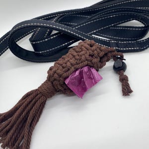 May include: A brown macrame dog waste bag holder with a tassel and a black leash with white stitching. The holder contains a purple waste bag. The leash is black with white stitching.