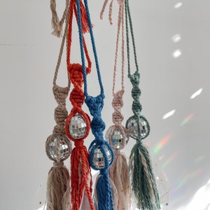 May include: Five colorful macrame tassel ornaments with mirrored disco balls hanging from hooks. The tassels are in shades of beige, red, blue, pink, and green.