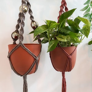 May include: Two macrame plant hangers with terracotta pots and a leafy plant. The hangers are made with gray and pink cord and feature decorative silver beads.
