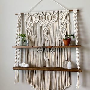 May include: A two-tiered wooden shelf with a macrame wall hanging. The shelf is made of dark wood and has a natural finish. The macrame hanging is made of white cotton cord and has a geometric pattern. There are small plants and decorative items on the shelves.