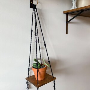 May include: A black macrame plant hanger with a wooden shelf and a small potted plant. The hanger is attached to a wall with a gold ring.