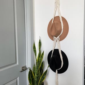 May include: A macrame hat rack with two hats hanging on it. The top hat is brown and the bottom hat is black. A potted snake plant is in the corner of the image.