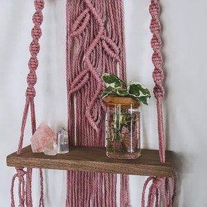 May include: A pink macrame wall hanging with a wooden dowel and fringe. A wooden shelf with a glass jar containing a plant and pink crystals is attached to the bottom of the hanging.