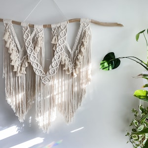 May include: A white macrame wall hanging with a natural wood branch at the top. The macrame is made with a variety of knots and has a fringe at the bottom. The hanging is in front of a white wall.