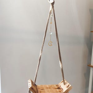 May include: A wooden hanging shelf with a triangular shape and a natural wood finish. The shelf is suspended by a thick rope with a decorative charm attached. The charm features a crescent moon and stars.