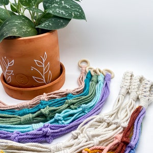 May include: A set of colorful macrame plant hangers with wooden rings. The hangers are made with different colored cords and are arranged in a rainbow pattern.