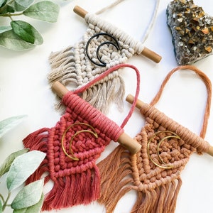 May include: Three macrame wall hangings with wooden dowels and fringe. The hangings are in cream, coral, and peach colors. Each hanging features a crescent moon shape with a gold or black metal hoop.