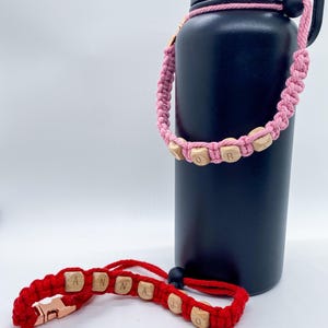 May include: A black insulated water bottle with a pink macrame strap featuring wooden letter beads spelling "DOR" and a red macrame wrist strap with wooden letter beads spelling "ANNA JO".