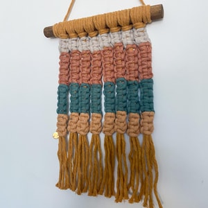 May include: A macrame wall hanging with a wooden dowel and hanging cord. The design features horizontal bands of color: mustard, off-white, coral, teal, and tan. The bottom has mustard-colored fringe and a small gold disc.