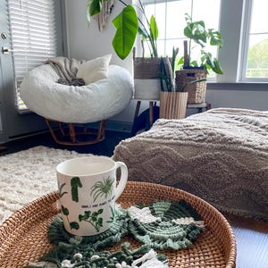 May include: A wicker tray holds a white mug with green plant illustrations and the text "Sorry, I have plants to see".  Green and white macrame coasters and a woven tray are on a light-colored rug. A fluffy white chair and plants are in the background.