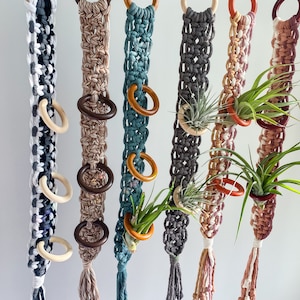 May include: Macrame plant hangers in various colors, including black and white, tan, teal, gray, and pink. Each hanger features wooden rings and a tassel at the bottom. Some hangers hold air plants in small terracotta pots.