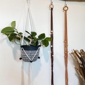 May include: Three macrame plant hangers with wooden beads. The left hanger is white with black beads and holds a black and white patterned pot with a plant. The middle hanger is brown with white beads. The right hanger is pink with gold beads.