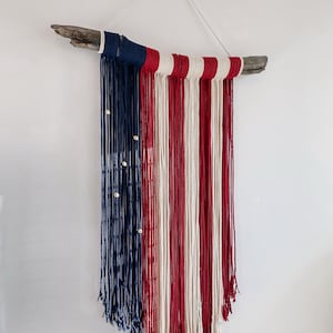May include: A red, white, and blue yarn wall hanging in the shape of an American flag. The yarn is strung on a piece of driftwood.