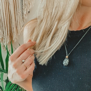 May include: A close-up of a woman wearing a black tank top and a necklace with a dark grey stone pendant. The necklace is made of a thin, dark grey cord.
