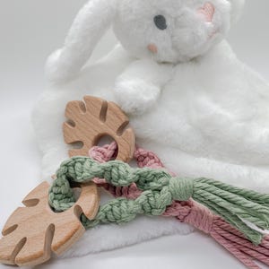 May include: A plush white bunny blanket with a pink nose and gray eyes. Two wooden leaf-shaped teethers are connected by a macrame cord in pink and green. A soft, white blanket is in the background.