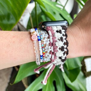 May include: A macrame Apple Watch band in white, brown, and mauve, paired with beaded bracelets. The bracelets feature clear, pink, and gold beads, with a letter "R" charm. The watch band is secured with a woven design.