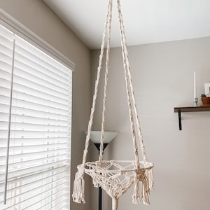 May include: A white macrame plant hanger with a circular top and a triangular base. The hanger has a long tassel hanging from the bottom.