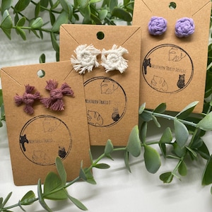 May include: Three pairs of handmade macrame stud earrings displayed on brown cardboard cards. One pair is mauve, one is off-white with frayed edges, and one is light purple. Each card features a circular logo with the text Northern Traced Designs.