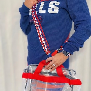 May include: Clear stadium tote bag with red handles and a red, white, and blue braided strap. The bag features a blue and red logo. The person is wearing a blue sweatshirt with the text "BUFFALO BILLS".