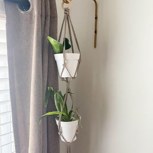 May include: Two white plant pots with green plants hanging from a macrame plant hanger. The hanger is attached to a gold metal hook on a white wall.